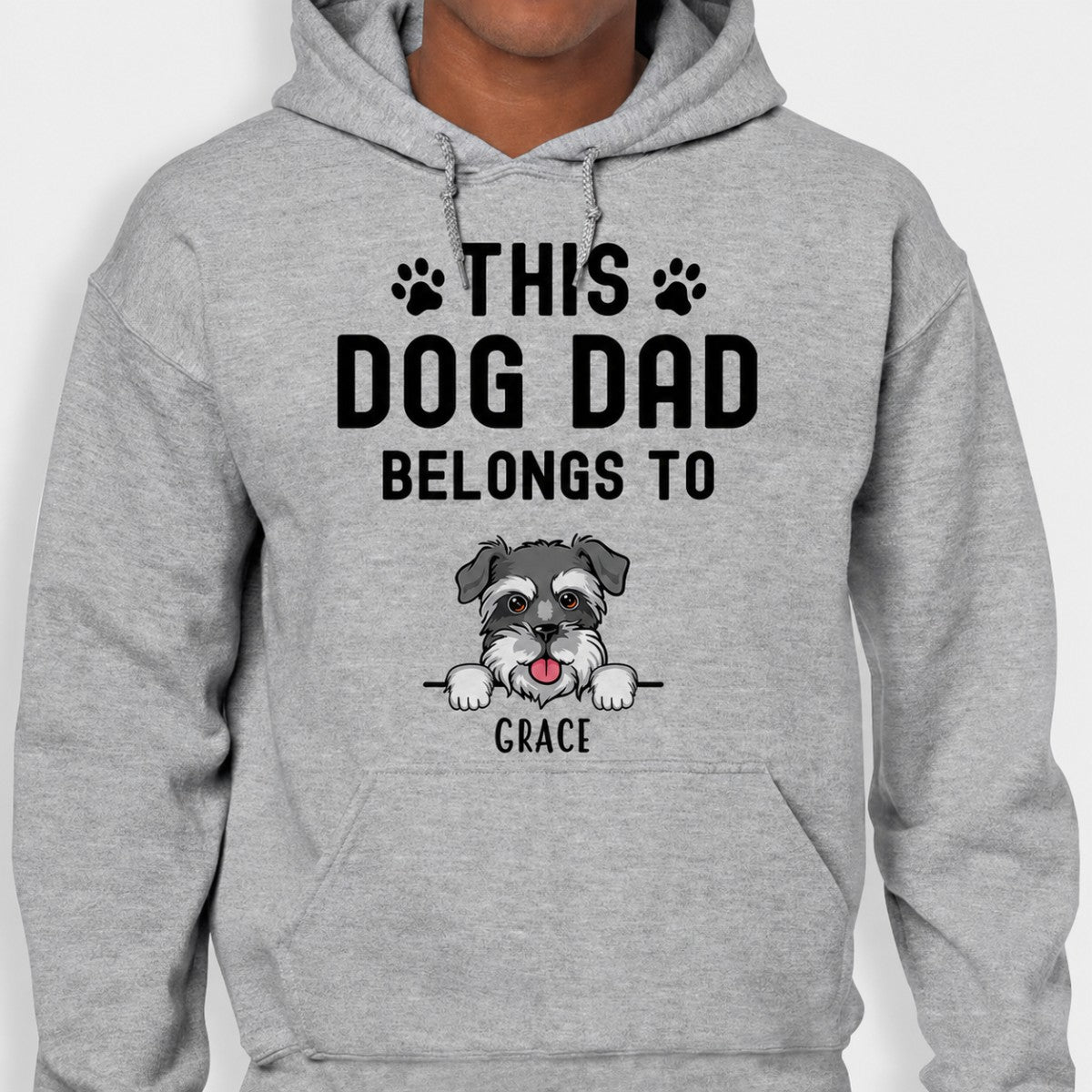 A person wearing a heather gray pullover hoodie with a black printed graphic that reads 'THIS DOG DAD BELONGS TO' above an illustration of a small gray and white dog with the name 'GRACE' below.